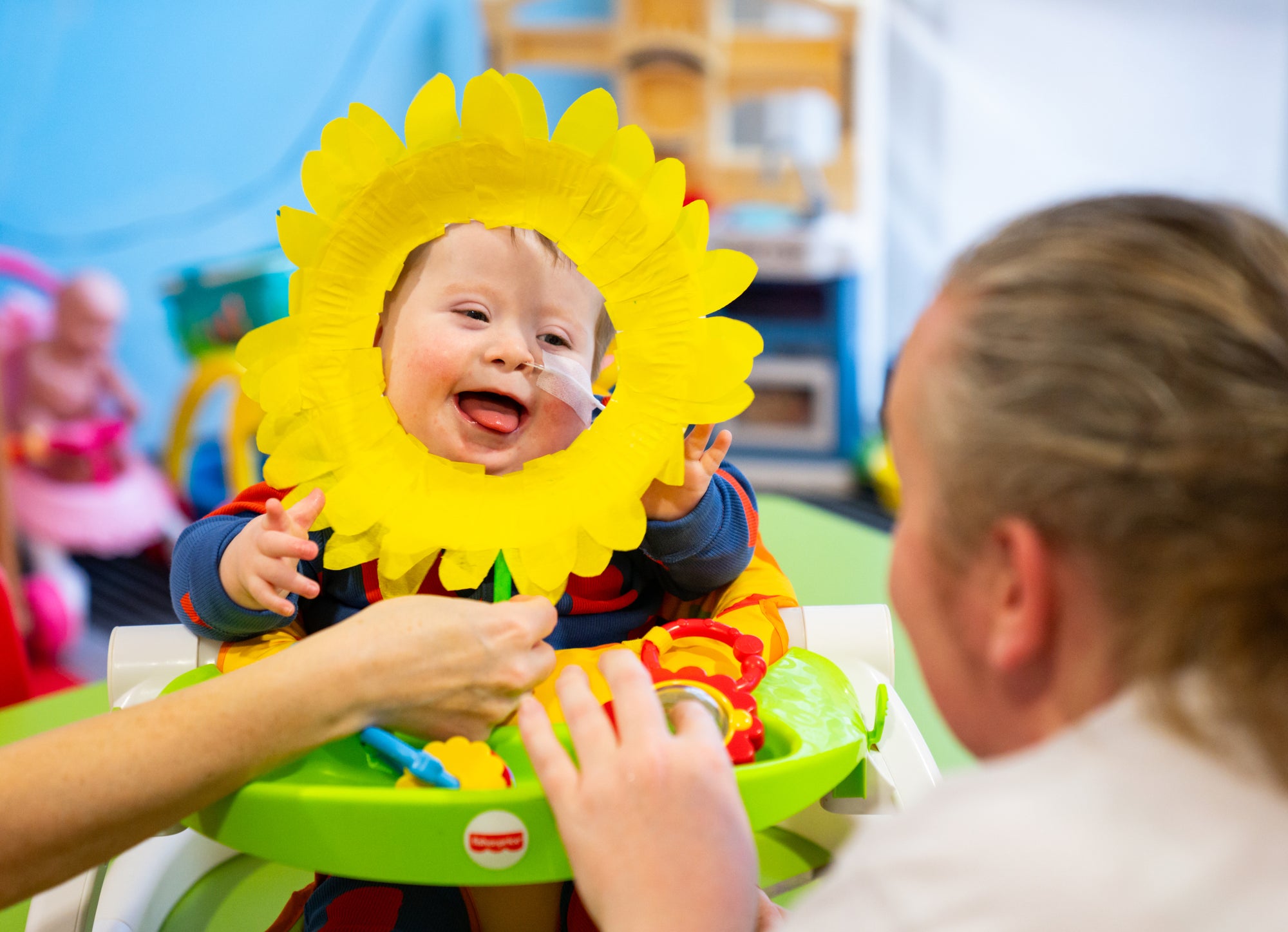 Child in a colorful baby seat with a sunflower headband, interacting with an adult in a playful setting.
